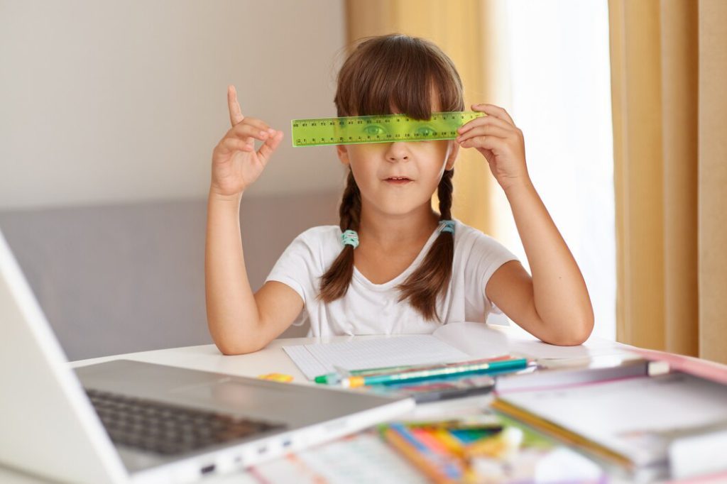 playful female child white t shirt sitting table front opened notebook covering eyes with green ruler distance education quarantine pointing finger up having idea 176532 15157
